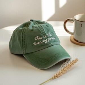 Op de afbeelding: Groene baseballcap met de geborduurde tekst "this is the turning point" in het wit. De cap is gemaakt van een zacht materiaal en heeft een gebogen klep. Een mok en een tarwe-aar staan op de achtergrond.
