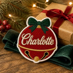 May include: A circular embroidered patch shaped like a red jingle bell with a dark green bow at the top. The name Charlotte is stitched in white cursive script across the centre. The patch features gold-coloured thread accents and a white border.