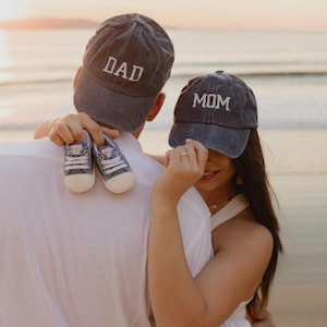 Puede incluir: Una pareja en una playa al atardecer, con gorras de béisbol de mezclilla a juego. Una gorra dice "DAD", la otra "MOM". La persona que lleva la gorra "DAD" sostiene un par de zapatillas de bebé azules. La pareja se abraza.