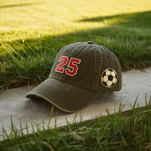 Puede incluir: Gorra de béisbol verde oliva con el número 25 en rojo y blanco. Un parche de balón de fútbol está en el lateral. La gorra está sobre una superficie de hormigón con hierba verde al fondo.