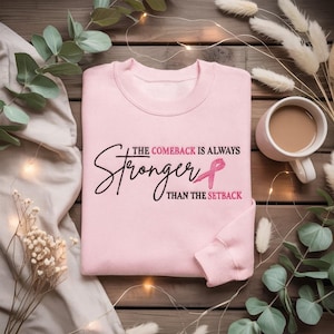 Könnte beinhalten: Hellrosa Sweatshirt mit dem Text "The Comeback is Always Stronger Than The Setback" und einer rosa Schleife. Das Sweatshirt liegt auf einer Holzoberfläche mit dekorativen Elementen und einer Tasse Kaffee.