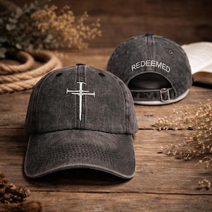 Puede incluir: Gorra de béisbol gris oscuro con una cruz blanca bordada en la parte delantera. La palabra "REDEEMED" está bordada en la parte trasera de la gorra. La gorra está hecha de una tela desgastada.