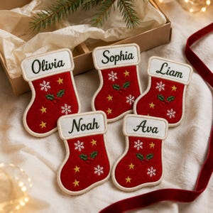 May include: Five red felt Christmas stocking ornaments with white cuffs embroidered with the names Olivia, Sophia, Liam, Noah, and Ava. Each stocking features embroidered white snowflakes, yellow stars, and green holly leaves with red berries.