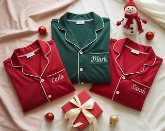 Puede incluir: Tres camisas de pijama personalizadas en rojo y verde con ribete blanco, cada una bordada con un nombre: "Carla", "Mark" y "Sarah". Una caja de regalo roja con una cinta crema, adornos navideños y un muñeco de nieve completan la escena festiva.