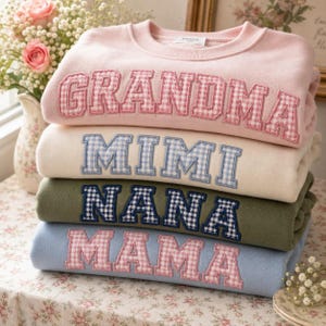 Könnte beinhalten: Ein Stapel pastellfarbener Sweatshirts mit gestickten Namen: "GRANDMA" in rosa Vichy, "MIMI" in blauem Vichy, "NANA" in marineblauem Vichy und "MAMA" in rosa Vichy. Die Sweatshirts sind auf einer Oberfläche mit floralem Muster gestapelt.