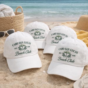 May include: Four white baseball caps featuring a dark green embroidered graphic of a beach umbrella and two lounge chairs with the text Cabo San Lucas Est. 2026 Beach Club. The caps are arranged on a sandy beach with the ocean in the background.