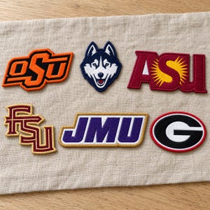 Puede incluir: Seis parches de tela bordados con varios logotipos universitarios y deportivos. Los parches incluyen el logotipo naranja de OSU, una cabeza de husky azul y blanca, el logotipo granate de ASU con un sol amarillo, el logotipo dorado de FSU, el logotipo morado de JMU y un logotipo G negro.