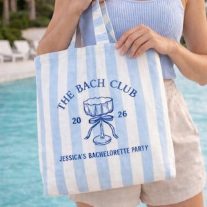 May include: A light blue and white striped tote bag with the text "THE BACH CLUB 2026" and "JESSICA'S BACHELORETTE PARTY" in blue. The bag features a drawing of a cocktail glass. The bag is held by a person.