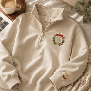 May include: A beige quarter-zip sweatshirt featuring an embroidered Christmas ornament design on the chest with a red bow, green wreath, and the letter A. The left sleeve cuff includes the embroidered name Anany above a small sprig of holly berries.