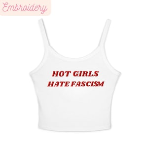 May include: White vest top with thin straps. The words "HOT GIRLS HATE FASCISM" are embroidered in red across the front. The top has a scoop neckline and is made of a soft, ribbed material. The word "Embroidery" is in the top left corner.