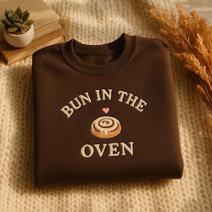 May include: A brown sweatshirt with the embroidered phrase "BUN IN THE OVEN" in white, featuring a heart and a cinnamon roll design. The sweatshirt is displayed on a cream-colored knitted surface.