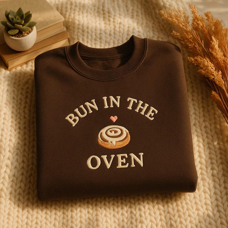May include: A brown sweatshirt with the embroidered phrase "BUN IN THE OVEN" in white, featuring a heart and a cinnamon roll design. The sweatshirt is displayed on a cream-colored knitted surface.