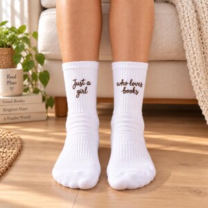 Könnte beinhalten: Ein Paar weiße Socken mit braunem Sticktext. Die linke Socke zeigt die Worte "Just a girl" und die rechte Socke die Worte "who loves books" in einer Schreibschrift. Die Socken haben eine gerippte Struktur am Schaft.