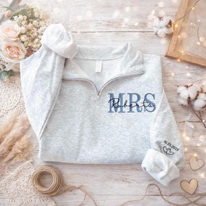 Puede incluir: Sudadera gris claro con cremallera de cuarto de círculo y texto bordado "MRS Roberts" en azul. El puño de la manga muestra la fecha "10.25.2025" y dos corazones. Prenda de vestir con temática nupcial o de boda.