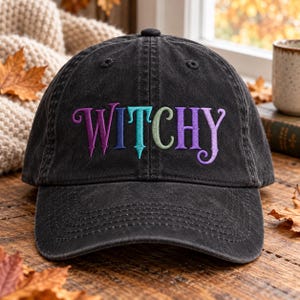 May include: A black baseball cap with a washed-out, vintage appearance. The word WITCHY is embroidered across the front in a stylised, decorative font. The letters feature a gradient of colours, transitioning from purple to blue, teal, and light green.