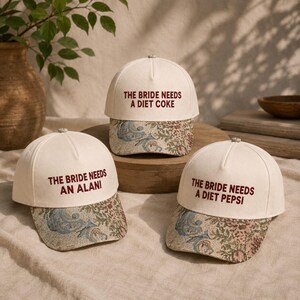 Puede incluir: Tres gorras de béisbol blancas con el texto burdeos "THE BRIDE NEEDS A DIET COKE", "THE BRIDE NEEDS AN ALANI" y "THE BRIDE NEEDS A DIET PEPSI". Las viseras presentan un estampado floral en tonos beige, azul y rosa.