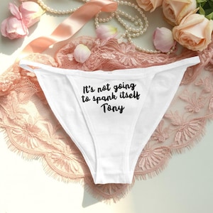 Könnte beinhalten: Weiße Unterwäsche mit dem Text "It's not going to spank itself Tony" in Schwarz. Die Unterwäsche wird auf einer rosa Spitzefläche mit Perlen, rosa Bändern und Rosen im Hintergrund präsentiert.