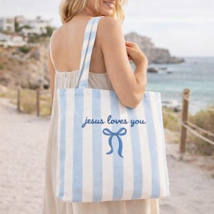 Puede incluir: Un bolso tote a rayas azules y blancas con el texto "jesus loves you" en fuente cursiva y un lazo azul. El bolso tiene asas a rayas azules y blancas a juego.