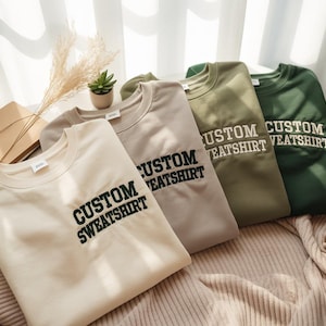 Pode incluir: Quatro sweatshirts personalizados em creme, taupe, verde oliva e verde floresta. Cada sweatshirt tem as palavras "CUSTOM SWEATSHIRT" bordadas em preto. Os sweatshirts estão dobrados e dispostos sobre uma superfície texturizada de tons neutros.