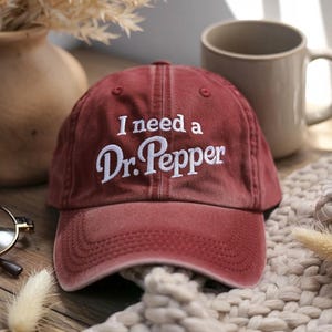 May include: A maroon baseball cap with the embroidered text "I need a Dr. Pepper" in white. The cap is sitting on a beige knitted surface, with a beige mug and a vase in the background.