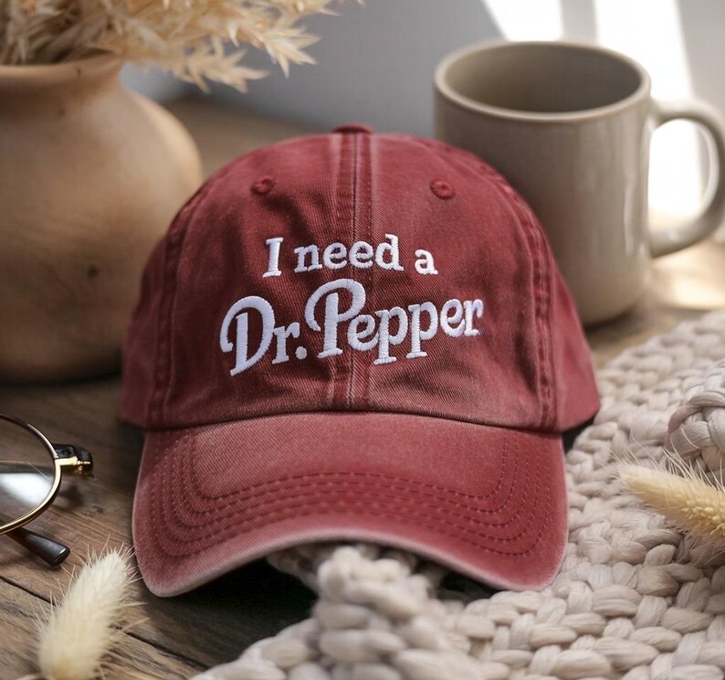 May include: A maroon baseball cap with the embroidered text "I need a Dr. Pepper" in white. The cap is sitting on a beige knitted surface, with a beige mug and a vase in the background.