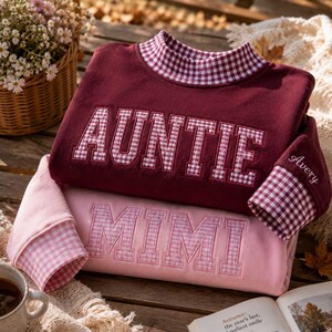 May include: A maroon sweatshirt with a high collar and cuffs featuring a red and white gingham pattern, embroidered with the word AUNTIE in gingham letters. A light pink sweatshirt with the word MIMI in pink gingham letters is layered below.