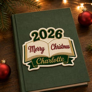 May include: A dark green hardback book featuring an embroidered patch on the front cover. The patch displays an open book graphic with the text 2026, Merry Christmas, and the name Charlotte in gold, red, and dark green thread colours.