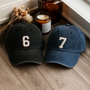 May include: Two baseball caps, one black and one navy blue, each with a white number stitched on the front. The black cap has the number 6, and the blue cap has the number 7. Both caps are on a wooden surface.