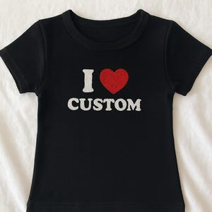 Peut inclure: T-shirt noir avec le texte "I ♥ CUSTOM" en blanc et un cœur rouge. Le t-shirt a des manches courtes et un col rond. Le tissu semble être côtelé.