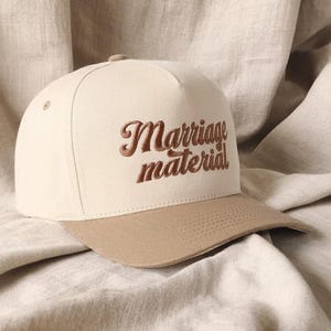 Könnte beinhalten: Beige Baseball Cap mit braunem Schirm. Auf der Kappe ist der Text "Marriage material" in brauner, kursiver Schrift gestickt. Der Hut ist vor einem neutralfarbenen Stoffhintergrund platziert.
