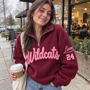 Könnte beinhalten: Ein kastanienbraunes Sweatshirt mit kurzem Reißverschluss, das auf der Brust das Wort Wildcats in erhabener, rosafarbener Schreibschrift zeigt, sowie den Namen Liam und die Zahl 24 in Rosa auf dem linken Ärmel, kombiniert mit einer cremefarbenen Stofftasche.
