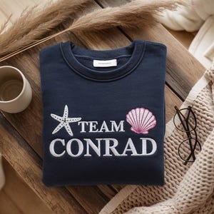 May include: Navy blue sweatshirt with white embroidered text "TEAM CONRAD", a starfish, and a pink seashell. A beige mug, eyeglasses, and a knitted blanket are also visible.