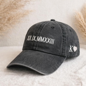 May include: A black baseball cap with white embroidered text and a heart symbol. The text on the front reads XII.IX.MMXXIII. The initials "K" and a heart are embroidered on the side. The cap is made of a soft, worn material.