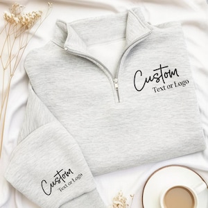 May include: A light grey quarter-zip sweatshirt featuring a silver metal zip at the collar. The fabric is printed with black text that reads Custom Text or Logo on the left chest and again on the sleeve cuff in a cursive and sans-serif font.