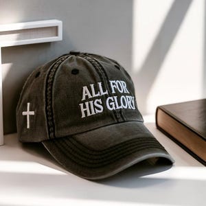 May include: A dark gray baseball cap with the embroidered text "ALL FOR HIS GLORY" in white. A white cross is embroidered on the side. The cap is sitting on a white surface next to a book and a white cross.