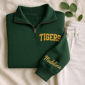 May include: A dark green sweatshirt with a quarter-zip collar and the word "TIGERS" embroidered in yellow. The sleeve features the name "Madeline" in yellow script. The sweatshirt is paired with white jeans and a sprig of greenery.