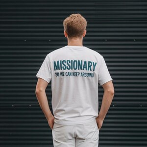 May include: White t-shirt with the word "MISSIONARY" printed in teal, followed by "SO WE CAN KEEP ARGUING" in a smaller font. The shirt is worn by a person standing in front of a dark, textured wall.