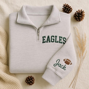 Könnte beinhalten: Ein hellgraues Sweatshirt mit einem Kragen mit Viertelreißverschluss. Das Wort "EAGLES" ist in Dunkelgrün auf die Brust gestickt. Der Ärmel zeigt den Namen "Jack" und eine Football-Grafik, ebenfalls in Grün. Zwei Tannenzapfen und ein Zweig trockenes Gras sind im Hintergrund.