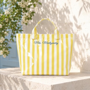 Peut inclure: Un sac fourre-tout à rayures jaunes et blanches avec une poignée. Le sac porte l'inscription "Mrs. Montgomery" en bleu. Le sac est posé sur une surface en pierre.