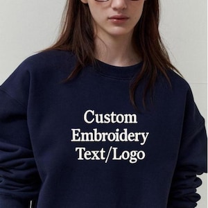 May include: Navy blue sweatshirt with the text "Custom Embroidery Text/Logo" in white. The sweatshirt is a crew neck style, with long sleeves. The image shows a close-up of the upper body.