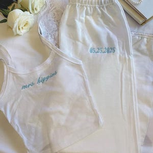 Puede incluir: Conjunto de pijama blanco con camiseta de tirantes y pantalón. La camiseta de tirantes tiene bordado "mrs higgins" en azul claro. El pantalón tiene bordado "05.25.2025" en azul claro. El conjunto está sobre una superficie blanca con rosas blancas.