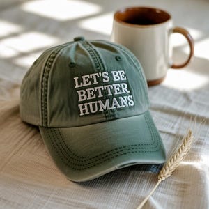 May include: An olive green baseball cap with the embroidered text "LET'S BE BETTER HUMANS" in white. A white and brown mug and a wheat stalk are in the background. The cap is made of a soft, worn material.