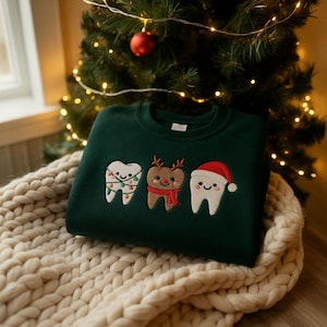 Könnte beinhalten: Dunkelgrünes Sweatshirt mit gestickten Zähnen im Weihnachtsstil. Ein Zahn ist mit Lichterketten geschmückt, ein anderer ist ein Rentier, und der dritte trägt einen Weihnachtsmannhut. Das Sweatshirt liegt auf einer cremefarbenen Strickdecke.