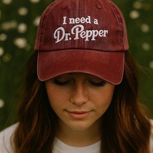 May include: A maroon baseball cap with the embroidered text "I need a Dr. Pepper" in white. The cap is worn by a person with long brown hair and a white shirt. The background is a blurred green and white.