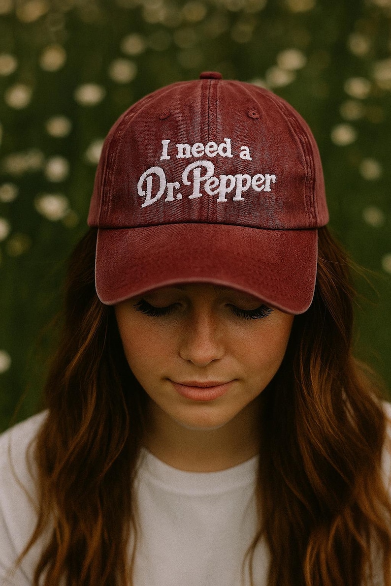May include: A maroon baseball cap with the embroidered text "I need a Dr. Pepper" in white. The cap is worn by a person with long brown hair and a white shirt. The background is a blurred green and white.