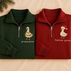Peut inclure: Deux sweat-shirts à fermeture éclair quart de cercle, un vert foncé et un rouge. Le sweat-shirt vert présente une oie brodée avec des lumières de Noël et les mots "grumpy goose". Le sweat-shirt rouge présente une oie portant un bonnet de Père Noël et les mots "festive goose".