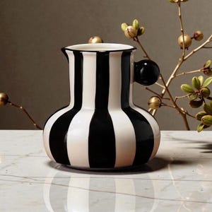Peut inclure: Une cruche en céramique à rayures noires et blanches avec une poignée ronde. La cruche a une base bulbeuse et un col étroit. La poignée est une sphère noire pleine. La cruche est posée sur une surface en marbre.