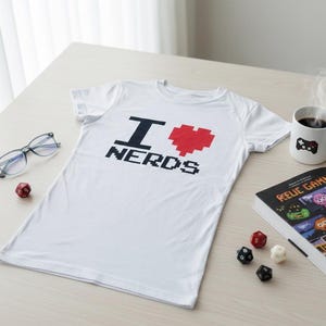 Op de afbeelding: Wit T-shirt met de tekst "I ♥ NERDS" in zwarte pixellettertype en een rood gepixeld hart. Het shirt ligt op een licht houten oppervlak, met een koffiemok, bril, dobbelstenen en een boek.