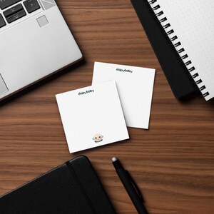 May include: Two white square notepads with the text "daipybaby" printed in teal. One notepad has a small cartoon illustration at the bottom. The notepads are on a wooden surface, with a laptop, notebook, and pen in the background.