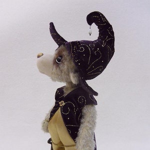 Könnte beinhalten: Ein Plüschhund, der einen lila-goldenen Narrenhut mit einer goldenen Taschenuhr am Bein trägt. Der Hund steht auf einem weißen Hintergrund.